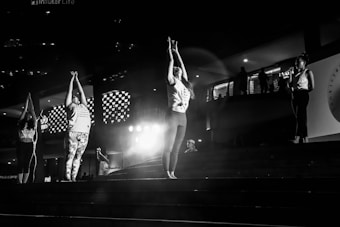 A group of people is practicing yoga or a similar activity on a set of steps in a dimly lit area. The central figures are stretching their arms upwards, while another person nearby is speaking into a microphone. The environment includes architectural elements such as patterned lights and railings, suggesting an urban or public space.