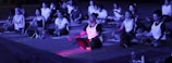 A group of people are sitting cross-legged on mats, engaging in meditation or a yoga session. The scene is likely outdoors, possibly in the evening due to the purple-blue lighting. Most individuals have closed eyes and are dressed in casual athletic wear, with some wearing printed shirts. Various personal items, such as bags and water bottles, are scattered around them.