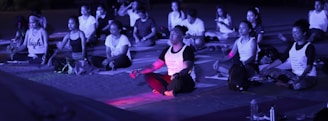A group of people are sitting cross-legged on mats, engaging in meditation or a yoga session. The scene is likely outdoors, possibly in the evening due to the purple-blue lighting. Most individuals have closed eyes and are dressed in casual athletic wear, with some wearing printed shirts. Various personal items, such as bags and water bottles, are scattered around them.