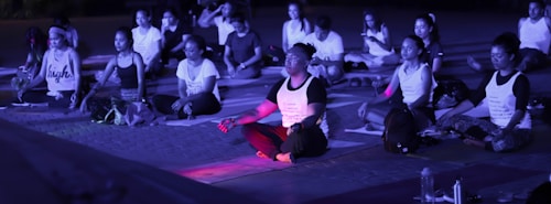 A group of people are sitting cross-legged on mats, engaging in meditation or a yoga session. The scene is likely outdoors, possibly in the evening due to the purple-blue lighting. Most individuals have closed eyes and are dressed in casual athletic wear, with some wearing printed shirts. Various personal items, such as bags and water bottles, are scattered around them.