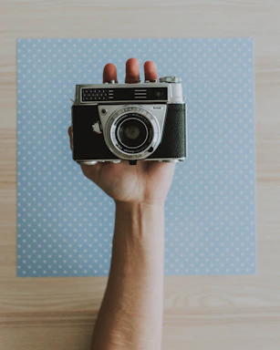 Close-up of a hand holding a vintage camera over a rustic wooden table with scattered food photography prints.