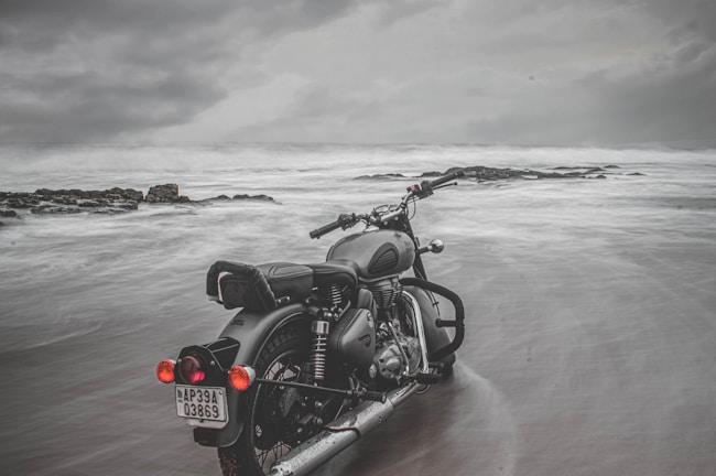 Motorbike parked on a coastal road in Europe with waves crashing nearby under a cloudy sky.