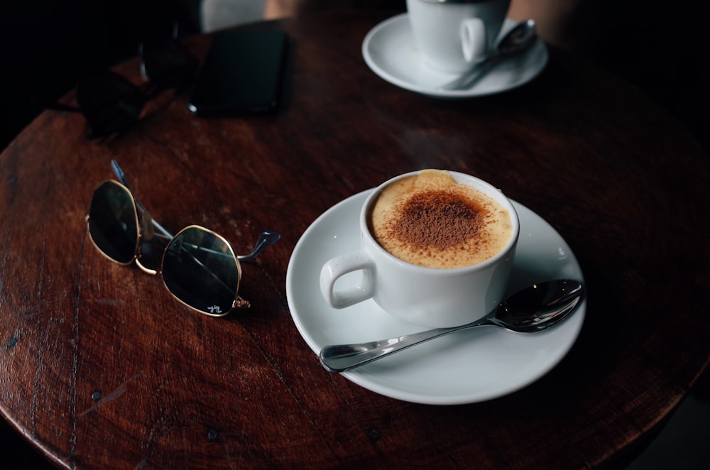 white ceramic cup with saucer beside silver framed aviator sunglasses