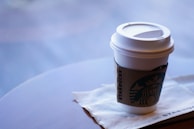 A white paper cup with a plastic lid features a cardboard sleeve displaying a logo. It is placed on a white napkin on a table with a blurred background.