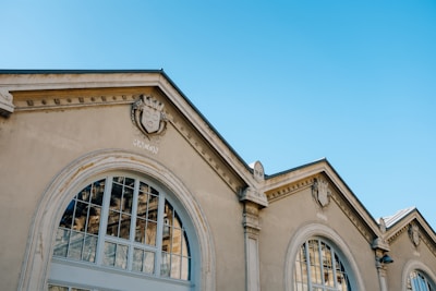 Facade of a historical building featuring large arched windows and detailed architectural elements, including decorative crest motifs on the gables. The building is made of light-colored stone and is set against a clear blue sky.
