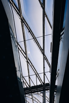 A modern architectural structure with a glass ceiling and metal frames. The ceiling allows natural light to filter through, creating a bright and airy atmosphere. The geometric lines of the framework add an industrial and contemporary aesthetic.