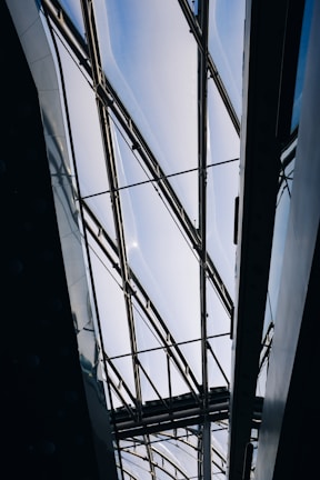 A modern architectural structure with a glass ceiling and metal frames. The ceiling allows natural light to filter through, creating a bright and airy atmosphere. The geometric lines of the framework add an industrial and contemporary aesthetic.