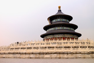 A traditional Chinese temple with a circular shape and three ornate, colorful tiers is surrounded by a white stone balustrade. This structure features intricate designs in blue, green, and gold, topped with a golden finial. The foreground consists of gray stone pavement leading to the temple, set against a pale, overcast sky.