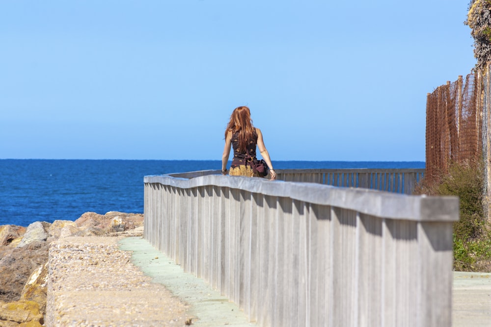 woman in brown jacket sitting on concrete fence near sea during daytime