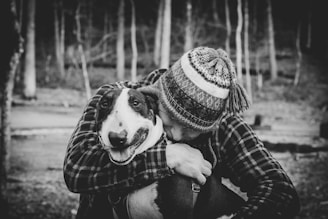 A black and white photograph depicts a person wearing a patterned winter hat and plaid jacket lovingly embracing a dog. The dog, a bull terrier, appears content with its tongue slightly out and eyes directed towards the camera. The background consists of defocused trees and a path in a forest setting, creating a tranquil atmosphere.