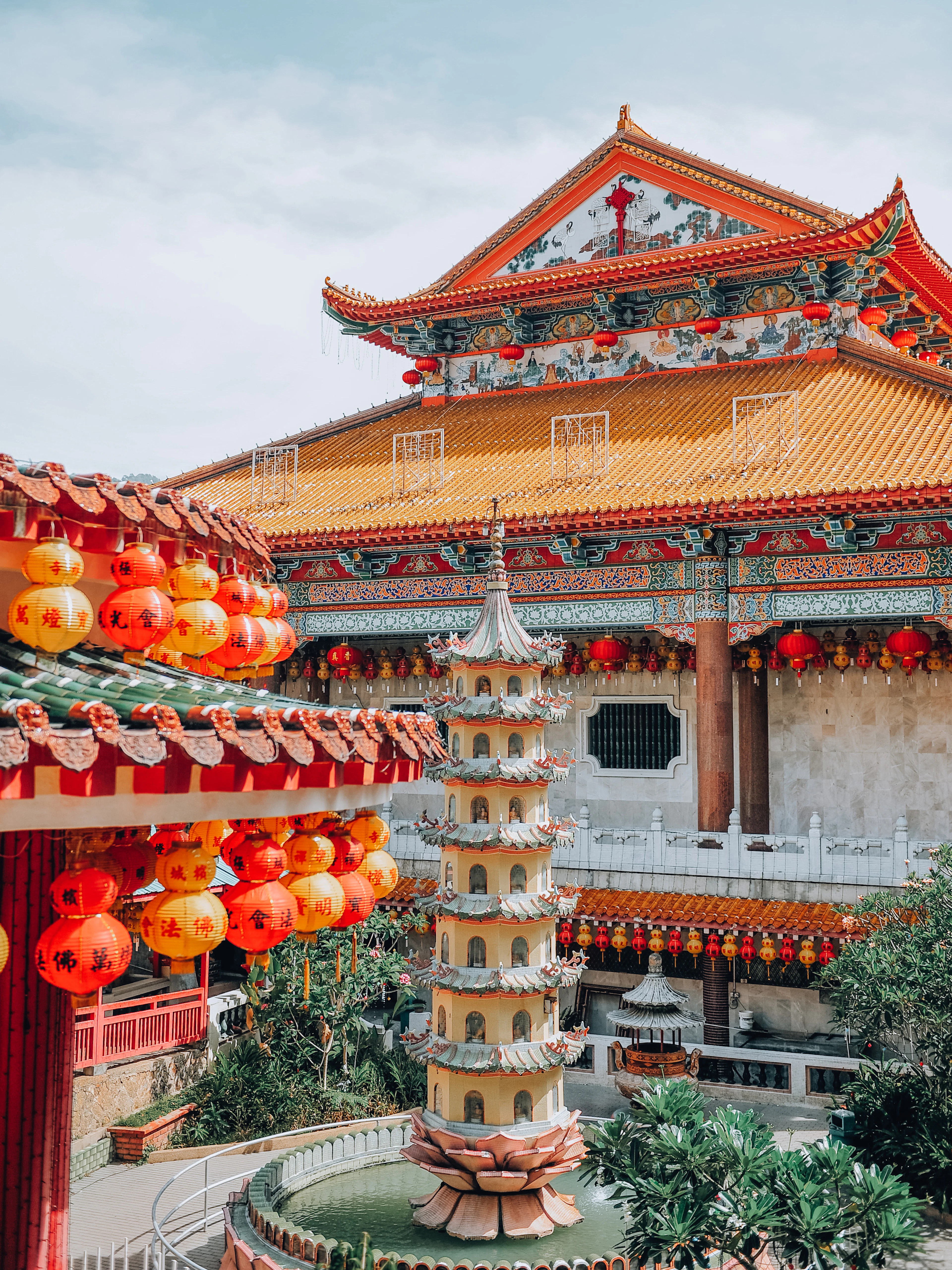 The ornate main hall of Kek Lok Si Temple in Penang, its orange tiled roof and painted eaves hung with cascades of red and yellow lanterns, a small tiered pagoda standing on a lotus base in the pond below.