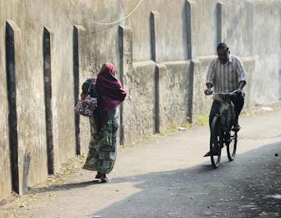 A woman dressed in traditional attire walks along a narrow lane beside a concrete wall, partly shaded by the soft sunlight. A man on a bicycle approaches from behind, creating a sense of daily routine in a quiet street.