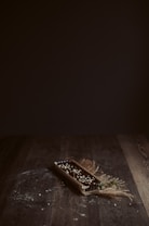 A rectangular tart is placed on a dark wooden surface, garnished with small white toppings and plant stalks. The lighting is dim, creating a warm, rustic ambiance, with scattered flour or crumbs surrounding the tart.