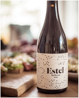 A bottle of wine labeled 'Estel Mallorca 2015' is placed prominently in the foreground. The label features a constellation motif with black dots and lines on a white background. Behind the bottle, there is a blurred, inviting spread of snacks or appetizers, set on a wooden board and partially visible, suggesting a casual dining or gathering atmosphere.