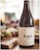 A bottle of wine labeled 'Estel Mallorca 2015' is placed prominently in the foreground. The label features a constellation motif with black dots and lines on a white background. Behind the bottle, there is a blurred, inviting spread of snacks or appetizers, set on a wooden board and partially visible, suggesting a casual dining or gathering atmosphere.