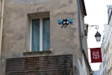 A beige, worn-out building exterior with a pixel art piece of a character featuring red eyes and blue wings on the wall. Below, there is a window with white frames and curtains. To the side, a traditional black street lamp hangs with a red sign featuring a white emblem.
