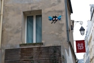 A beige, worn-out building exterior with a pixel art piece of a character featuring red eyes and blue wings on the wall. Below, there is a window with white frames and curtains. To the side, a traditional black street lamp hangs with a red sign featuring a white emblem.