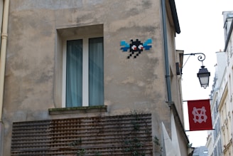A beige, worn-out building exterior with a pixel art piece of a character featuring red eyes and blue wings on the wall. Below, there is a window with white frames and curtains. To the side, a traditional black street lamp hangs with a red sign featuring a white emblem.