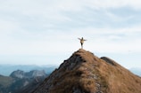 A man wearing a rugged jacket stands atop a mountain ridge, overlooking a vast green valley under a clear blue sky.