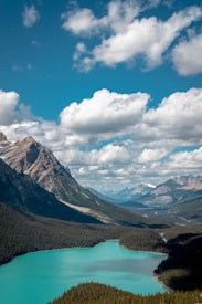 A stunning landscape featuring a turquoise-colored lake surrounded by dense forests and majestic mountains. The sky is vibrant blue, dotted with fluffy white clouds. The scene conveys a sense of natural beauty and tranquility, with the rugged peaks contrasting the smooth surface of the water.