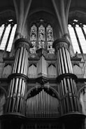 A large and ornate church pipe organ with multiple tall pipes arranged in a symmetrical pattern. The organ is positioned against a backdrop of intricate stained glass windows that allow light to filter through, illuminating the space with a serene atmosphere. The architectural details include arches and columns, emphasizing the grandeur of the setting.