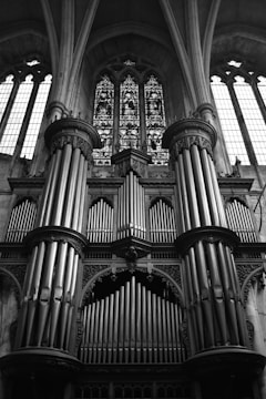 A large and ornate church pipe organ with multiple tall pipes arranged in a symmetrical pattern. The organ is positioned against a backdrop of intricate stained glass windows that allow light to filter through, illuminating the space with a serene atmosphere. The architectural details include arches and columns, emphasizing the grandeur of the setting.