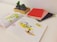 An open book is spread on a white table, showing colorful illustrations and diagrams. Next to it is a wooden tray with two small potted cacti. To the right of the book, there are three closed notebooks in red, pink, and dark purple, each clipped with gold paper clips.