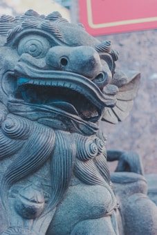 A close-up view of a traditional stone sculpture depicting a mythical creature with intricate carvings, including detailed fur and an open mouth showing teeth. The creature has an expressive facial expression, large eyes, and a mane curling around its head.