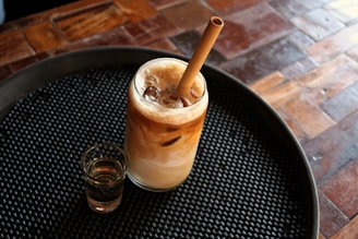 A glass filled with an iced coffee beverage featuring a frothy layer on top is placed on a round, textured black serving tray. Alongside the glass, there is a small shot glass containing a light-colored liquid. The setting includes a wooden table with various shades and grains, adding warmth to the scene.