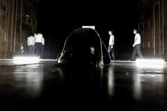 A dimly lit stage with several people on it, some walking and others stationary. The foreground features a person bent over in the center, surrounded by bright floor lights that cast dramatic shadows.