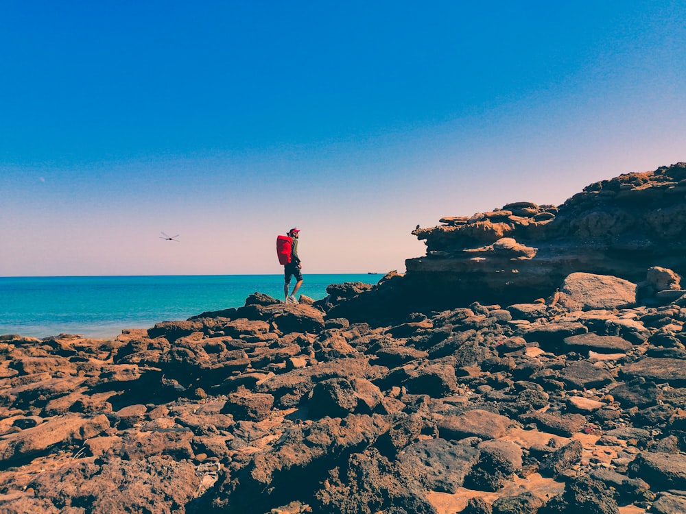 man in red shirt standing on rock formation near sea during daytime