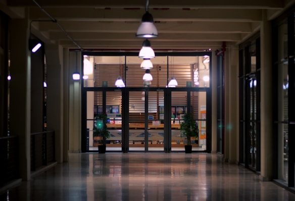 A long hallway with polished floors leading to a set of glass doors. The doors reveal a warmly lit room inside, featuring hanging pendant lights and several potted plants. The space beyond the doors appears to be an office or reception area with brochures and office equipment visible.