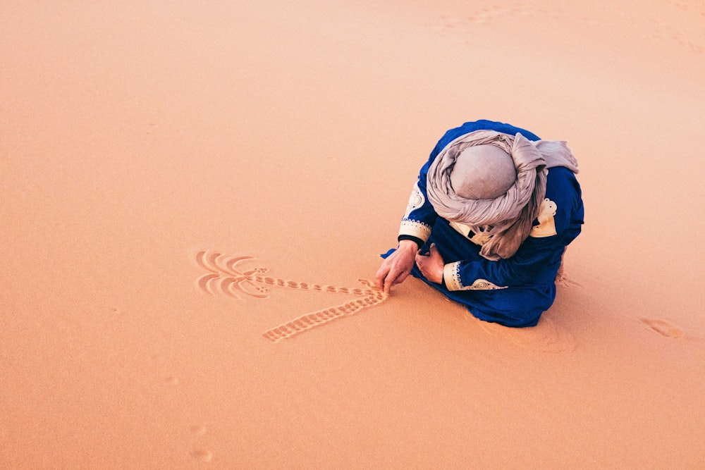 man in blue jacket and blue denim jeans sitting on brown sand during daytime