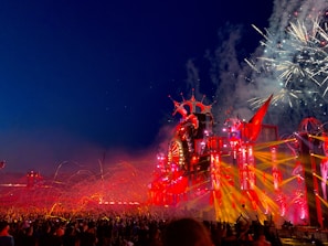 An energetic outdoor festival at night with a large crowd gathered in front of an elaborate stage adorned with vibrant red and orange lights. Fireworks explode in the night sky, adding to the lively atmosphere.