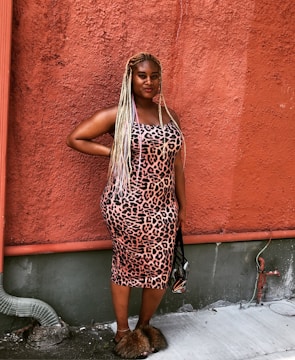 A person stands confidently against a textured, reddish wall wearing a leopard print dress and holding a small handbag. Their long, braided hair has a striking appearance and they are wearing fluffy sandals. A small patch of concrete and a metal pipe are visible at the bottom of the image.