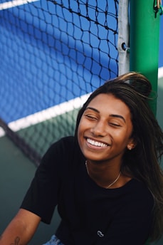 A person is smiling broadly while sitting next to a tennis court net. The background shows a blue tennis court, and the person appears to be relaxed and joyful.