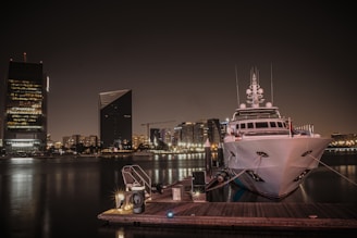 An elegant luxury yacht docked at a vibrant marina with city skyline in the background.