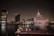 A large luxury yacht docked at a marina during nighttime, with the city skyline illuminated in the background. The reflection of the lights in the water creates a serene and elegant atmosphere. The yacht's sleek design and the modern buildings behind it are clearly visible.