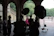 A group of people gathers under an ornate arched structure, engaged in a photoshoot. A woman in a vivid pink dress is in focus, illuminated by large lighting equipment. The background features ornate stonework and lush green trees, with a few people walking in the distance.