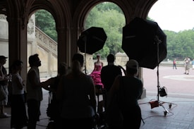 A group of people gathers under an ornate arched structure, engaged in a photoshoot. A woman in a vivid pink dress is in focus, illuminated by large lighting equipment. The background features ornate stonework and lush green trees, with a few people walking in the distance.