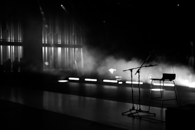 A dimly lit stage setup with a prominent microphone stand and a stool. There is a mist or smoke effect in the background, creating a moody ambiance. Vertical light strips along the back wall add to the dramatic atmosphere.