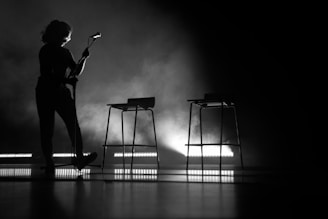 A silhouetted figure stands on stage holding a microphone, while two high stools are set in the background against a dramatic backdrop of smoke and backlighting.