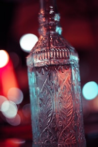 A tall, intricately designed glass bottle with ornamental patterns is in focus. The background is blurred, featuring vibrant bokeh lights in warm red and cool blue tones.