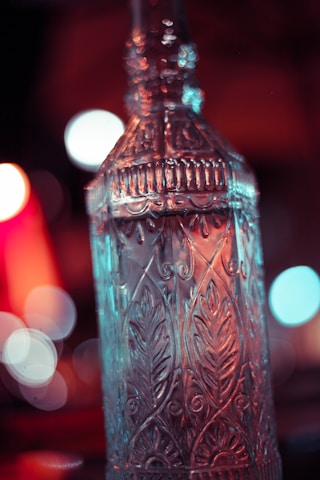 A tall, intricately designed glass bottle with ornamental patterns is in focus. The background is blurred, featuring vibrant bokeh lights in warm red and cool blue tones.