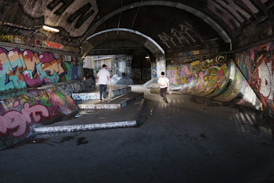 A dimly lit urban skate park situated under a bridge, with extensive graffiti covering the walls. Two people are skateboarding, engaging in tricks on the ramps and steps. The area is marked by a mix of vibrant street art and shadowed lighting.