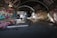 A dimly lit urban skate park situated under a bridge, with extensive graffiti covering the walls. Two people are skateboarding, engaging in tricks on the ramps and steps. The area is marked by a mix of vibrant street art and shadowed lighting.