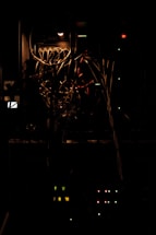 A dimly lit server rack with tangled cables and lit indicator lights. The atmosphere is dark, and the light source is minimal, emphasizing the complex web of networking wires. The lights in the rack are a combination of green and red, with some yellow, indicating the operational status of various components.