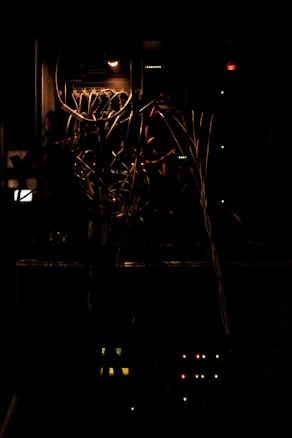 A dimly lit server rack with tangled cables and lit indicator lights. The atmosphere is dark, and the light source is minimal, emphasizing the complex web of networking wires. The lights in the rack are a combination of green and red, with some yellow, indicating the operational status of various components.