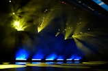 Bright yellow stage lights pierce through smoke in a dark setting, creating a dramatic and vibrant atmosphere. The beams of light spread across the stage, illuminating the area with touches of blue light at the bottom.