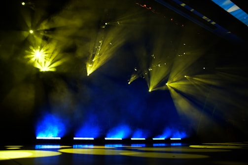 Bright yellow stage lights pierce through smoke in a dark setting, creating a dramatic and vibrant atmosphere. The beams of light spread across the stage, illuminating the area with touches of blue light at the bottom.